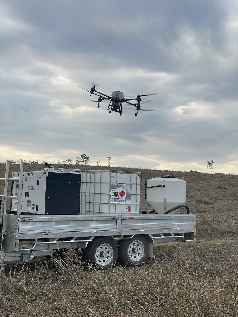 A spreading drone in the air above a trailer carrying a generator, a product tank and a hopper.