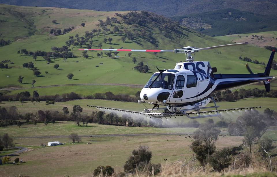 A helicopter with a spray boom flying over green hill country.